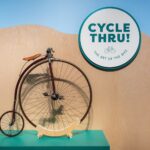 Penny-farthing bicycle in a museum exhibit with a round sign reading "Cycle Thru! The Art of the Bike"