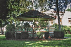 Bandstand with peaked shingled roof surrrouned by short hedges and trees in a grassy park