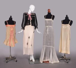 Group with 3 partial manneqins with slip dresses/nightgowns in different lengths and shades of pink, an 1 full mannequin wearing long slip with a few scattered flowers and a sheer black jacket with a pink bow
