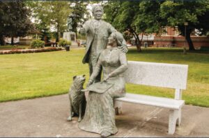 Statue of a standming man, seated woman on a park bench with a book on her lap, and dog sittting on the ground