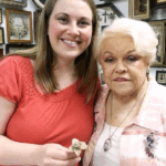 A smiling young woman and white-haired older woman hold handmade flowers in front of a background of framed art
