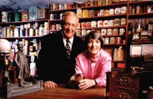A tall man in a dark suit and shorter woman in a pink blouse stand smiling behind a desk with a display of antique cans behind them