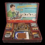 Open box with "Atomic Energy Lab" and a picture of a boy on the interior lid, toy parts and instruction books in compartments