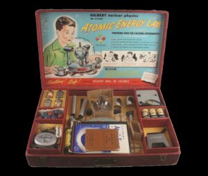 Open box with "Atomic Energy Lab" and a picture of a boy on the interior lid, toy parts and instruction books in compartments