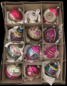 Open cardboard box with 12 shiny Christmas ornaments in pink, teal, and silver tones with stripes, flowers, and glitter